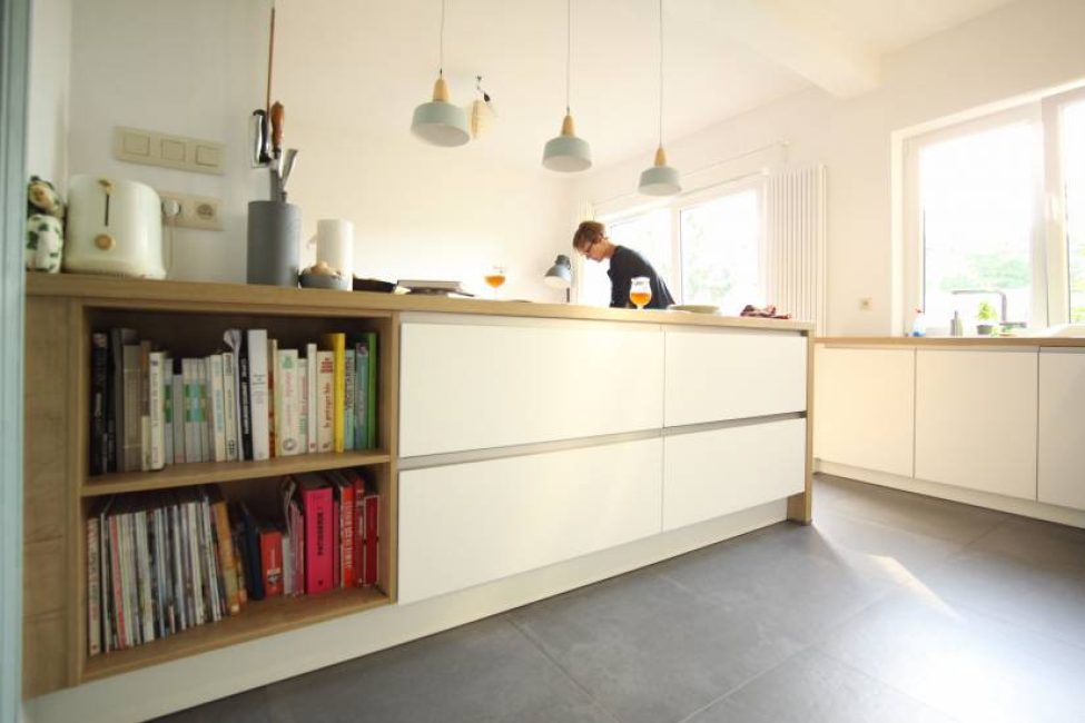 Cuisine moderne avec îlot central blanc, étagère de livres intégrée, et suspensions lumineuses grises. Une personne se tient près de la fenêtre.