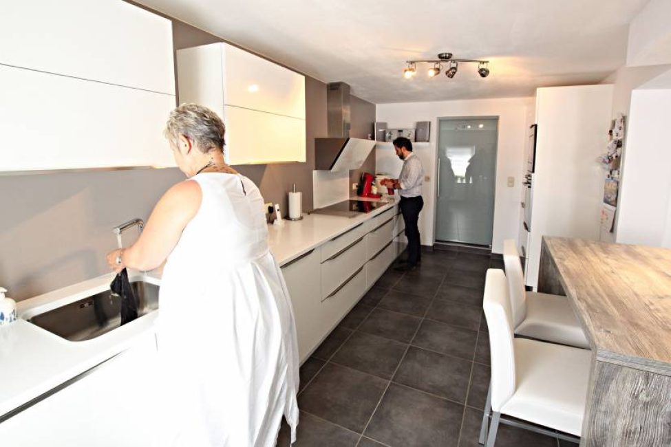 Cuisine moderne avec des placards blancs, un plan de travail clair, une femme au lavabo et un homme près d'une machine à café.