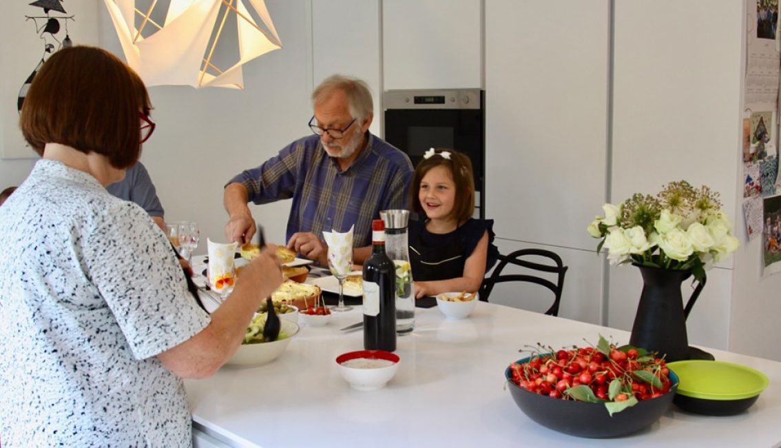 Trois personnes autour d'une table blanche avec des plats, des verres de vin, une carafe d'eau, un vase de fleurs blanches et un bol de cerises.