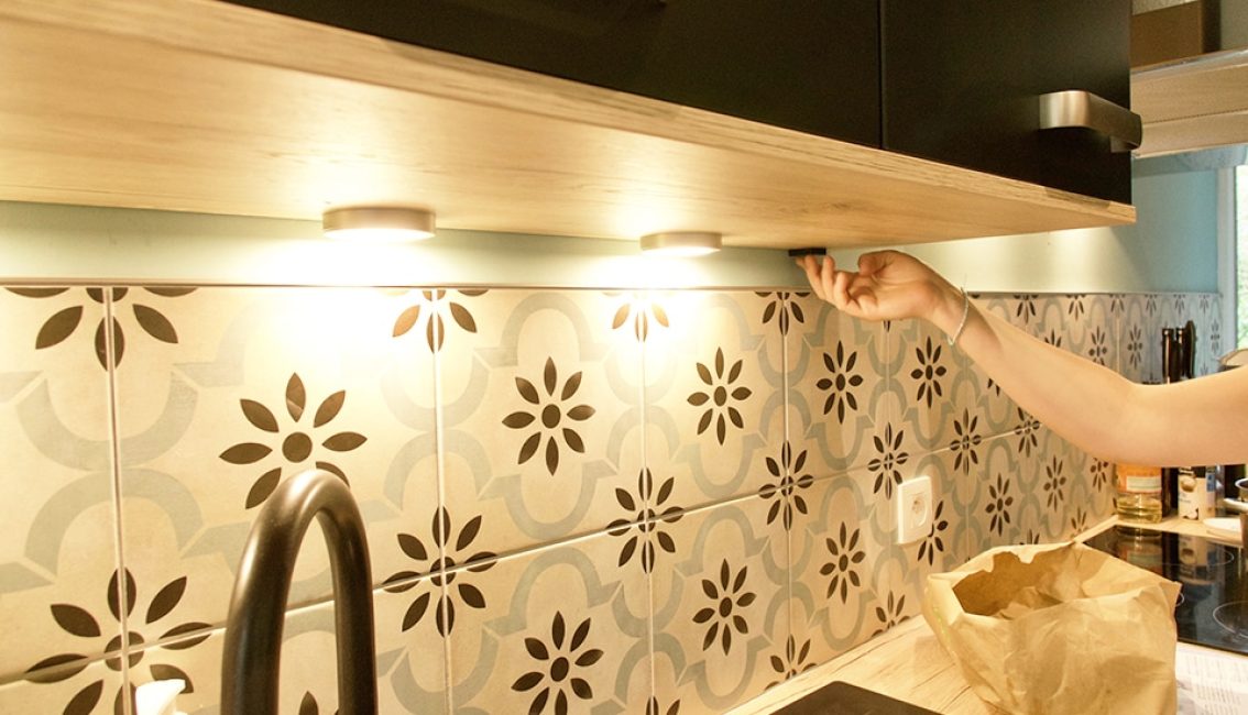 Une main allume un éclairage sous un meuble de cuisine, avec un dosseret en carreaux à motifs géométriques et un évier noir.