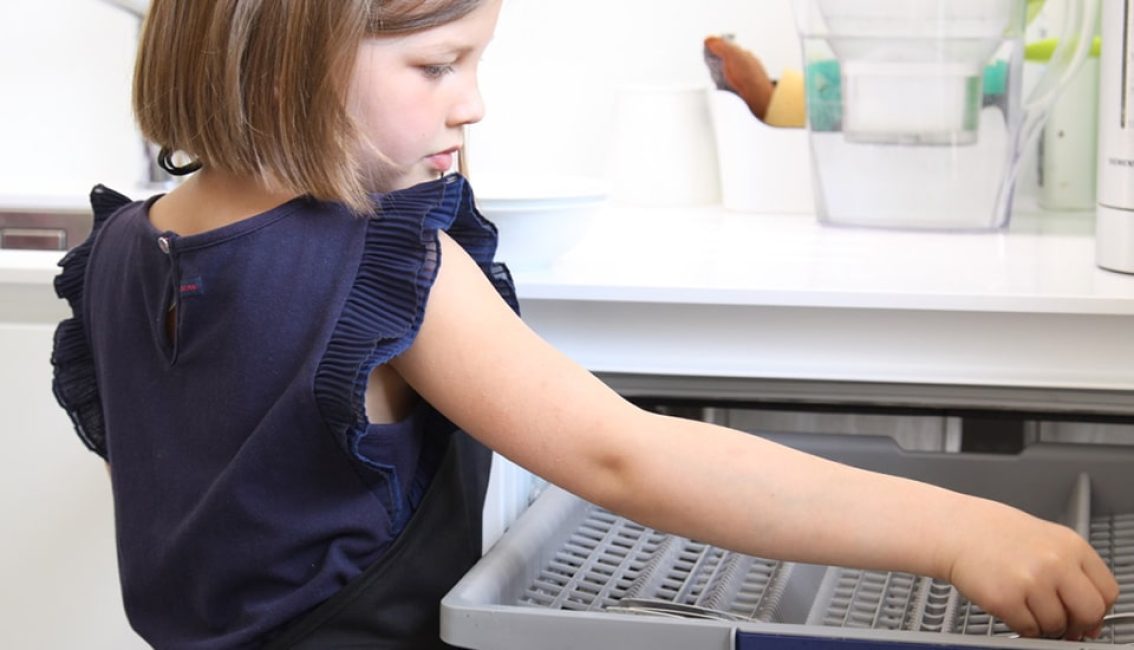 Une enfant en haut bleu ouvre le tiroir d'un lave-vaisselle dans une cuisine blanche avec une carafe filtrante sur le comptoir.