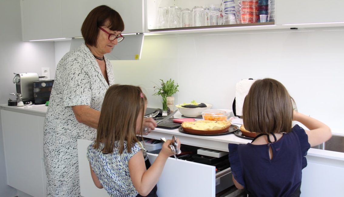 Une femme et deux enfants préparent des plats dans une cuisine moderne blanche avec un plan de travail garni de tartes.
