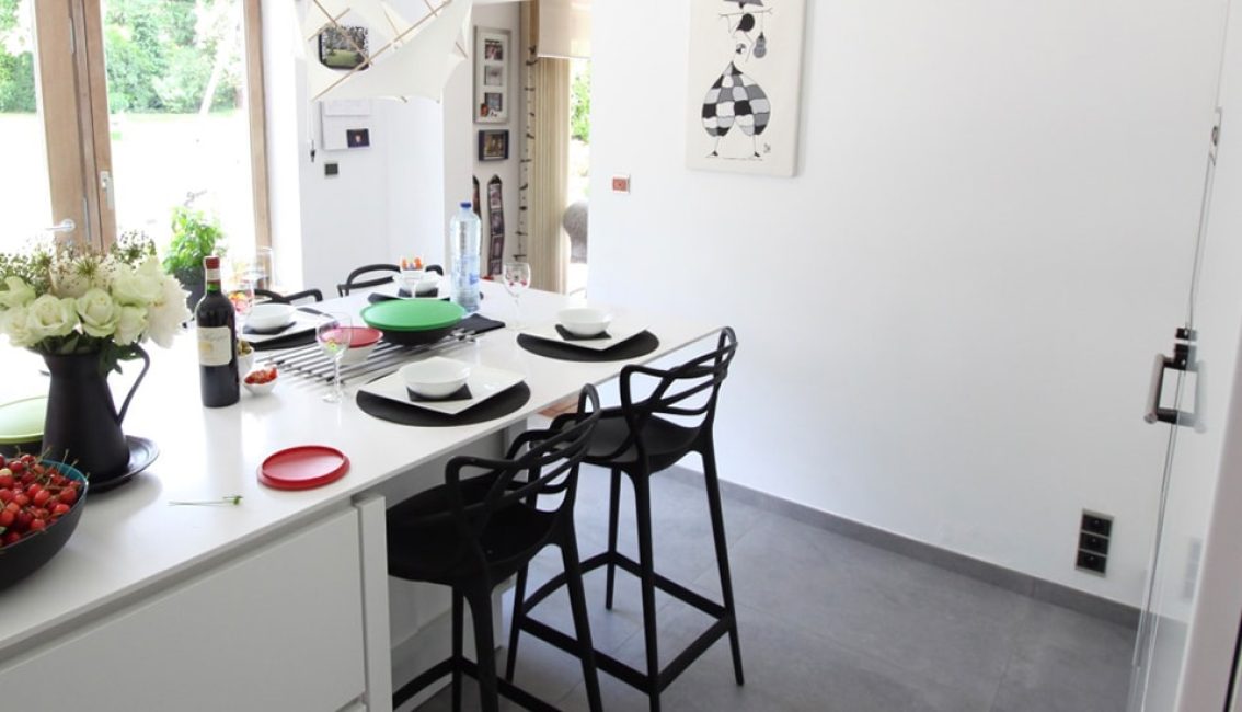 Cuisine moderne avec un îlot central blanc, des chaises noires, des assiettes noires et blanches, et un vase de fleurs blanches.