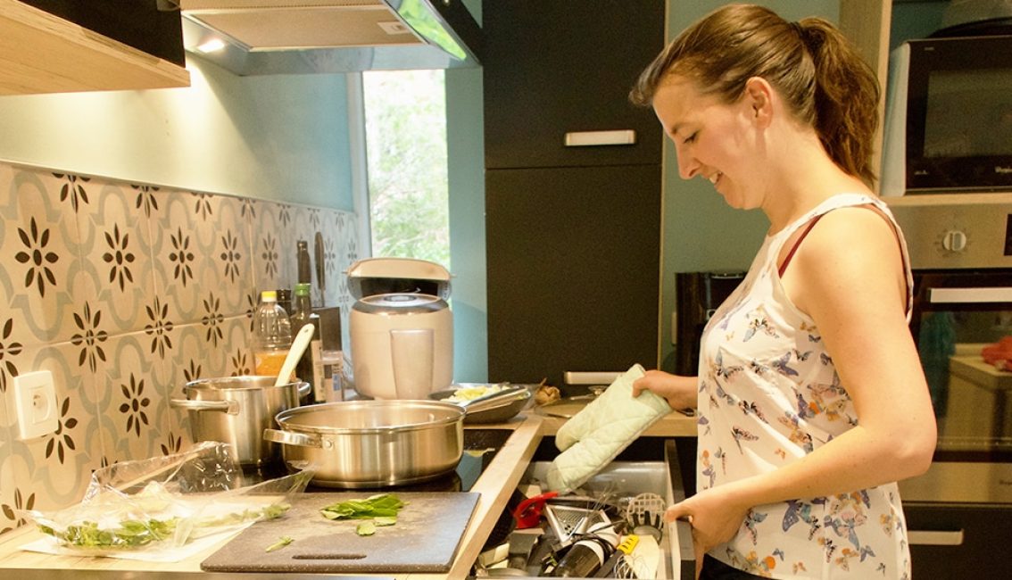 Une femme cuisine dans une cuisine moderne avec des casseroles en acier inoxydable et un robot culinaire sur le plan de travail.