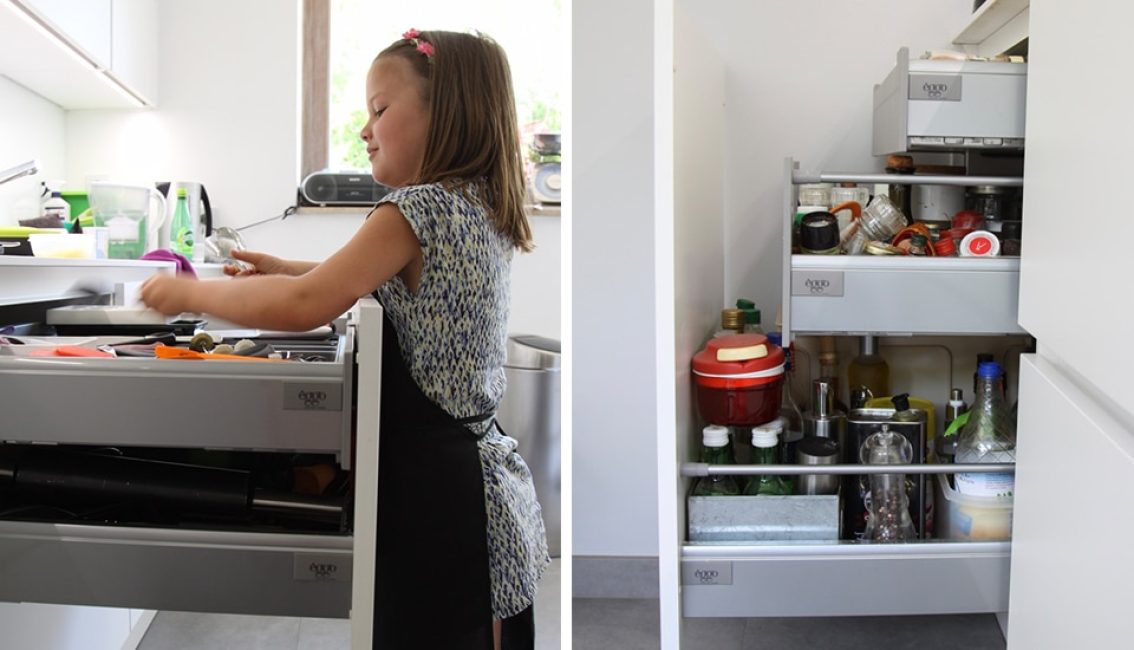 Une fillette organise des ustensiles de cuisine dans un tiroir gris, à côté d'un placard rempli de bouteilles et condiments.