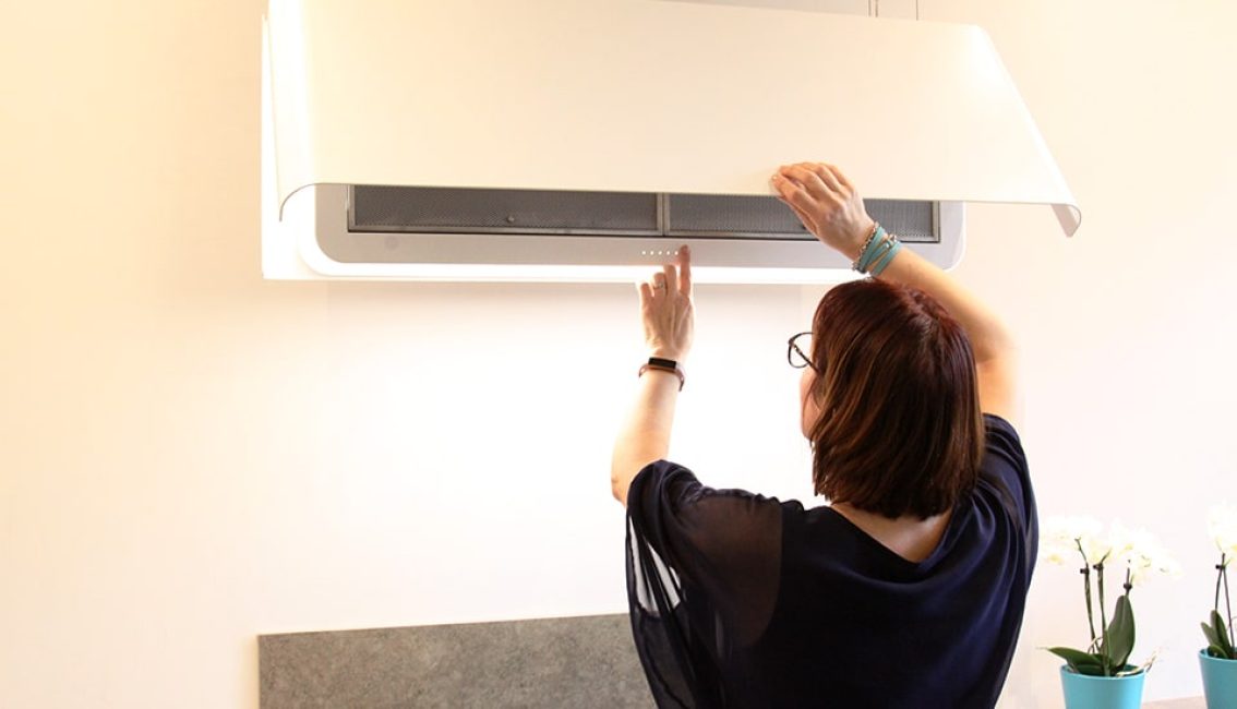 Une personne ajuste une hotte de cuisine blanche, des fleurs blanches dans un vase turquoise sont visibles à droite.