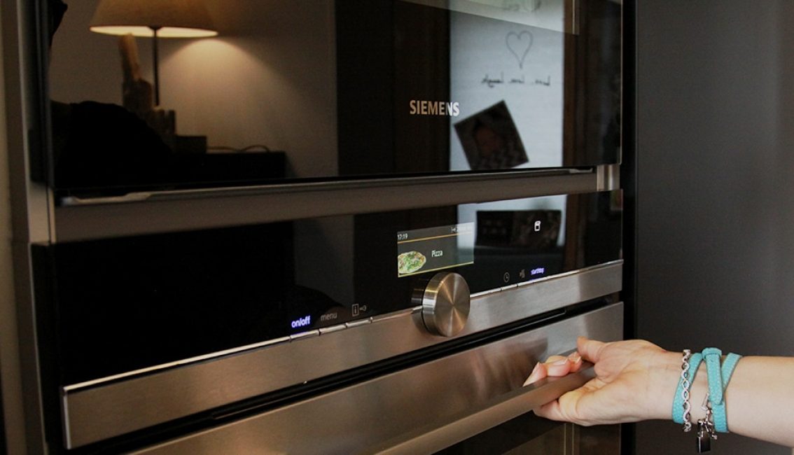 Une main ouvre un four encastré Siemens noir et argent, avec un écran affichant "Pizza", dans une cuisine moderne.