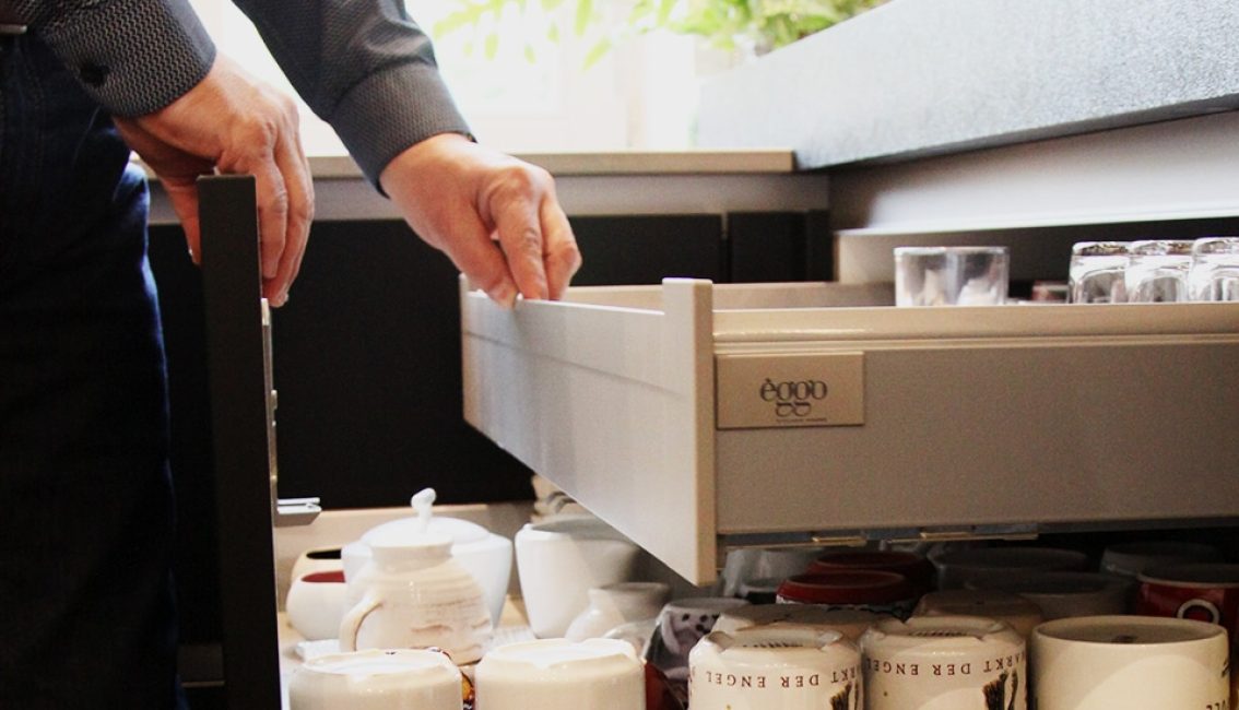 Une personne ouvre un tiroir de cuisine gris contenant des verres et des tasses blanches et rouges, avec une théière à proximité.