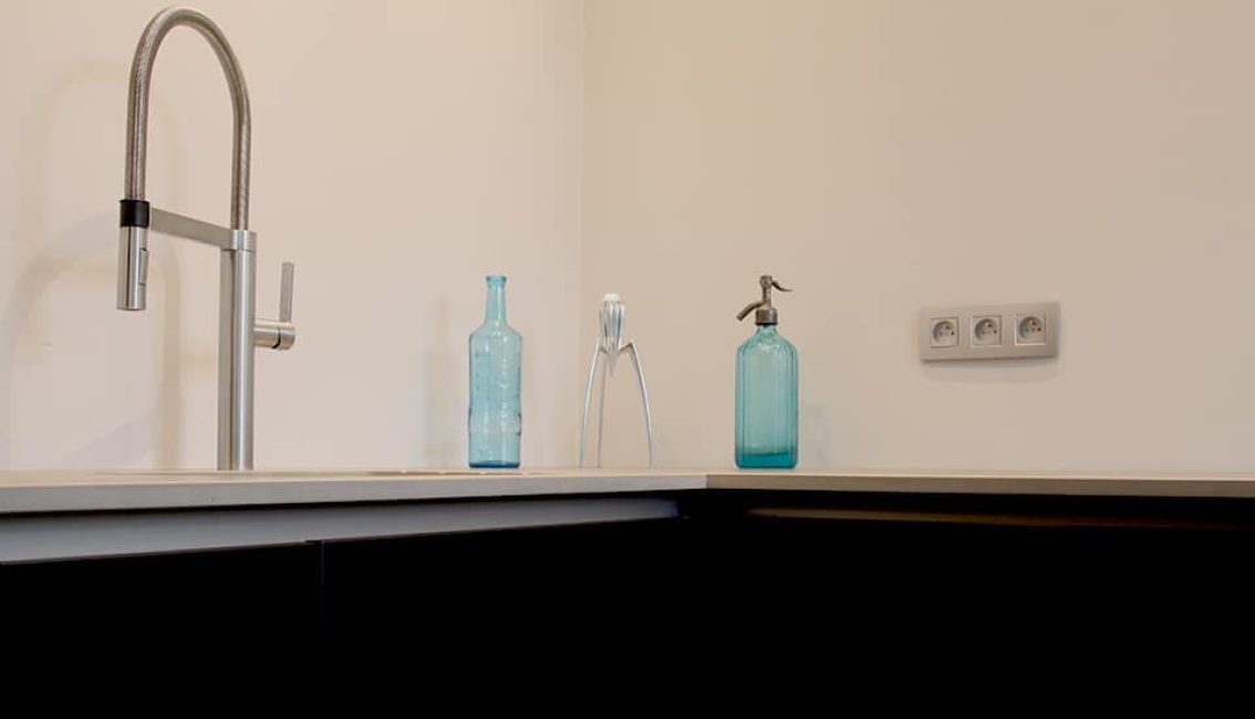 Un robinet en métal, deux bouteilles bleues et un presse-agrumes argenté sont disposés sur un plan de travail beige avec des prises électriques murales.