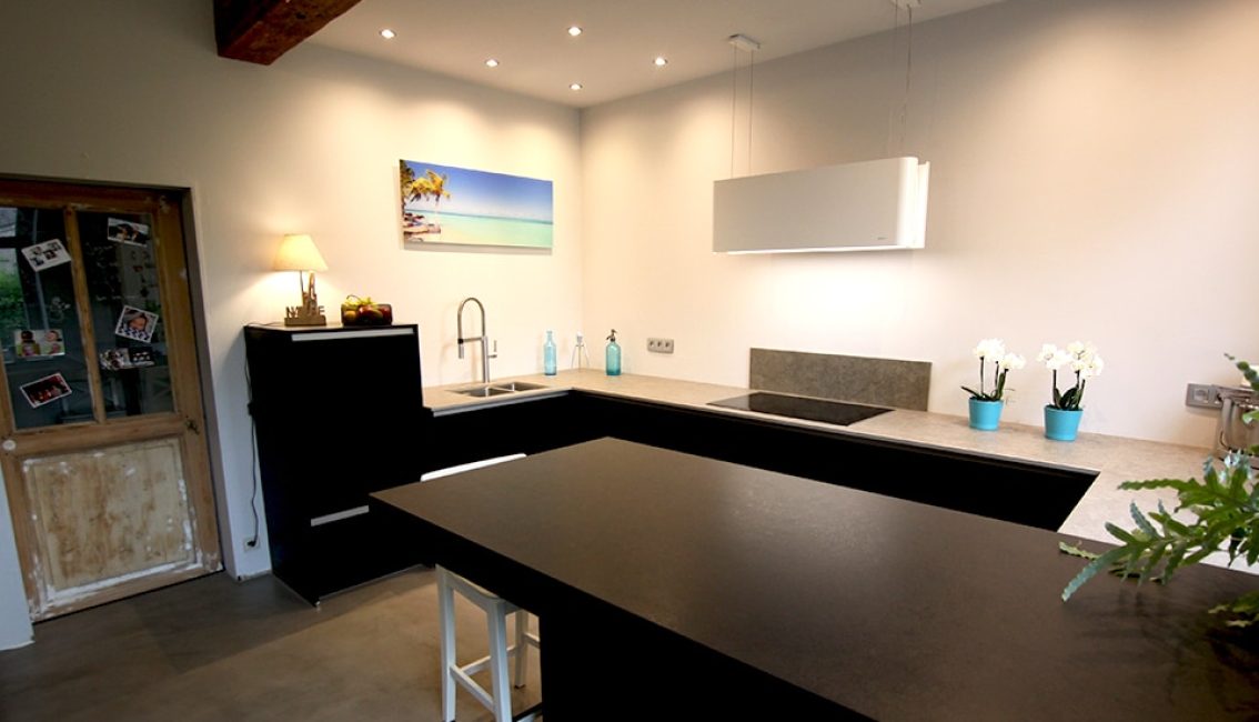 Cuisine moderne avec comptoirs noirs, mur blanc, tableau de plage, trois plantes en pots bleus et éclairage encastré.