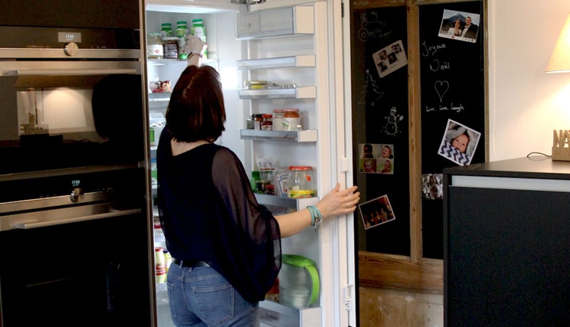 Une femme en jean et chemisier noir ouvre un réfrigérateur rempli de produits alimentaires. À côté, un tableau noir affiche des photos et des dessins.