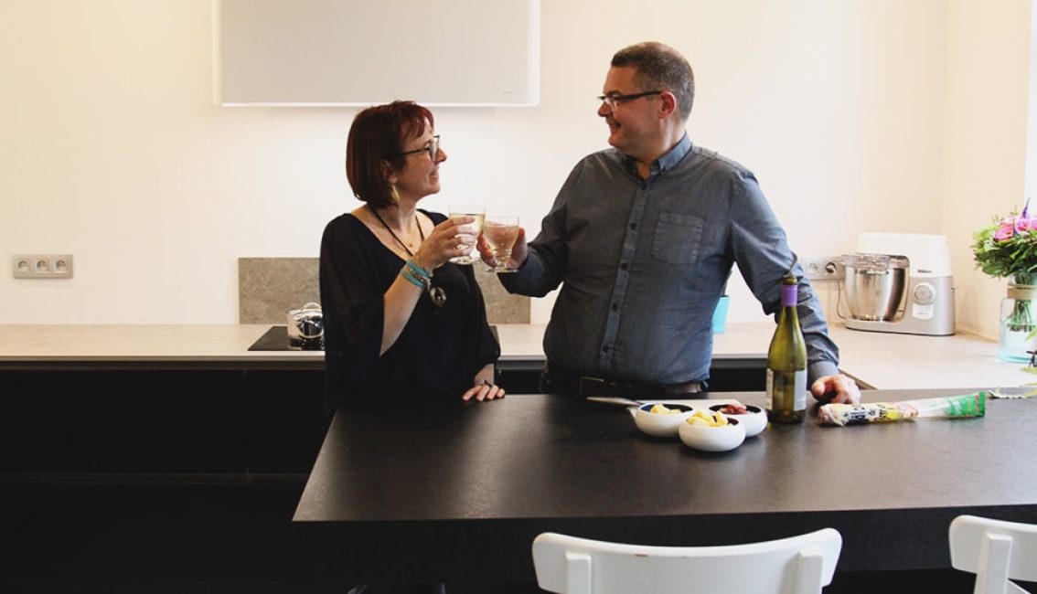 Deux personnes trinquent dans une cuisine moderne avec un plan de travail noir, des bols de nourriture et une bouteille de vin.